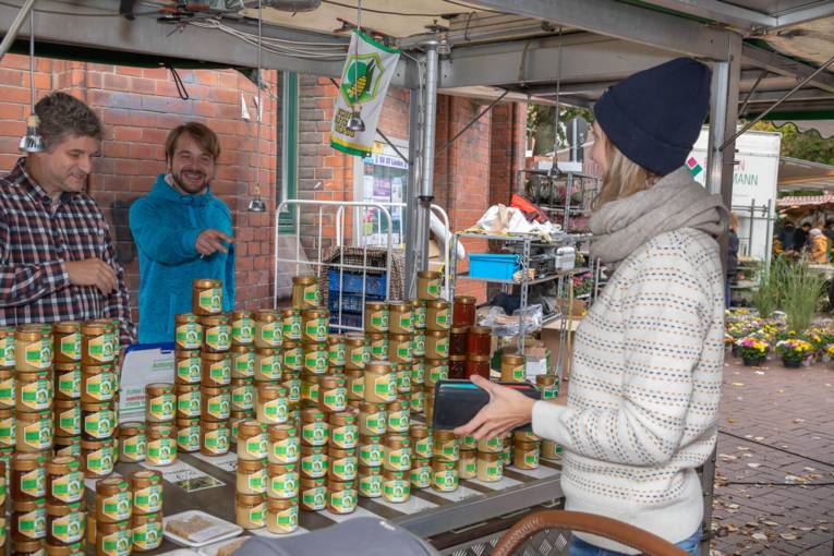 Zwei M&auml;nner bieten einer Kundin Honig an.
